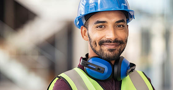 Portrait of satisfied construction site manager wearing safety vest and blue helmet with copy space. Young middle eastern architect watching construction site with confidence and looking at camera.