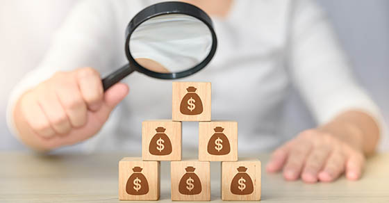 search for money. hand holding magnifying glass investigating wooden block with money bag icons.  money management concept. passive income. money saving concept.