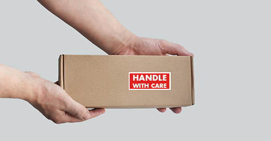 One rectangular cardboard box is held in the hands on a plain light background. A box with an adhesive warning label for packaging - Fragile, Handle with care, Thank you. No people. Corner composition