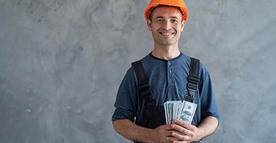 a happy worker in a protective helmet received a salary of American dollars and holds them in his hands, stands near a concrete wall. Construction of the payday. Payment for the work of the foreman